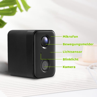 Schwarze kompakte Überwachungskamera mit zwei Linsen und grünen Beschriftungen (Mikrofon, Bewegungsmelder, Lichtsensor, Blinklicht, Kamera) auf hellem Hintergrund